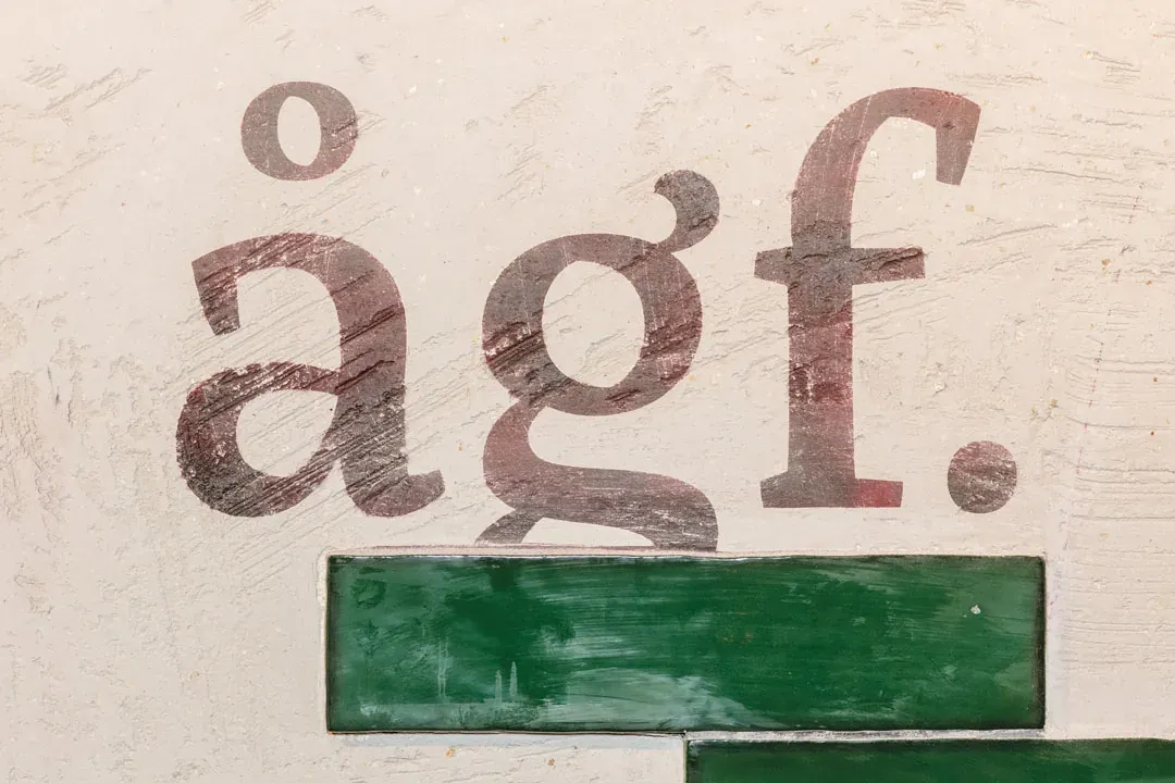 Der Schriftzug ågf. in dunklem Rot auf dem Kalkputz, darunter eine Reihe grüner Fliesen