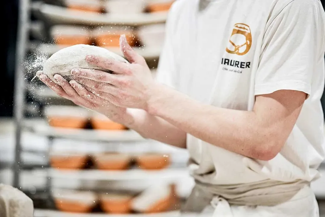 Zwei Hände formen einen Brotteig, im Hintergrund Brotlaibe im Gärkorb-Regal
