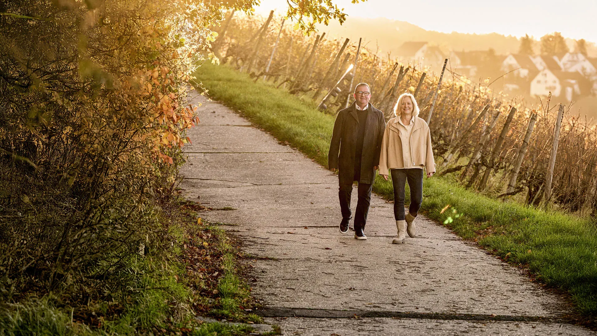 Tobias Maurer und seine Frau Claudia auf einem Weg durch die Weinberge im Herbst, dahinter im Gegenlicht die Häuser der Stadt