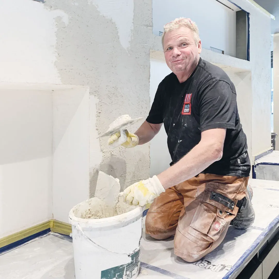 Kurt Schief kniet vor der Wand und zieht mit der Kelle Kalkputz auf, daneben der Eimer mit der Masse