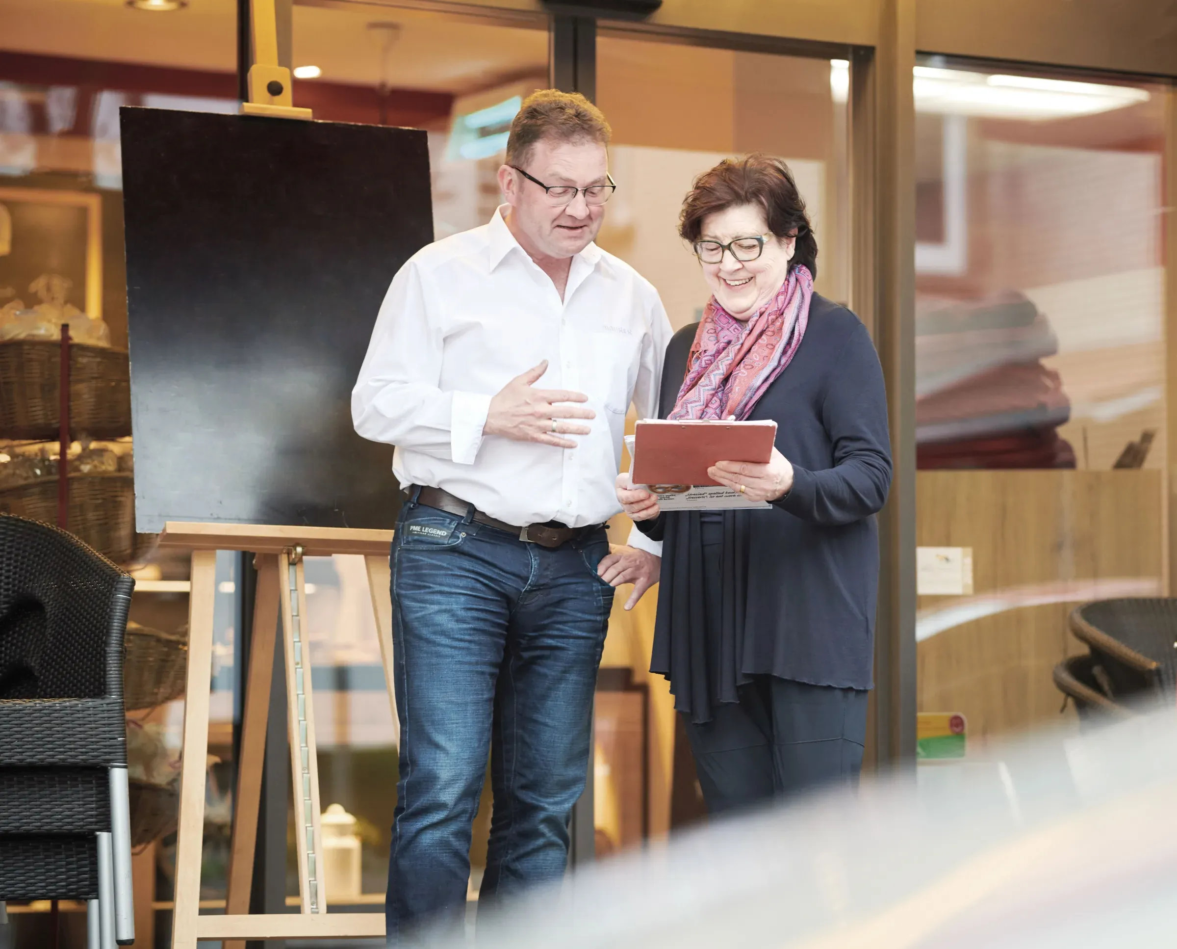 Tobias Maurer und seine Mutter Anne Maurer vor einem Bäckerei-Café, gemeinsam über eine Liste gebeugt