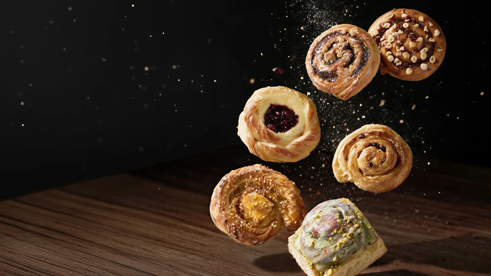 Sechs Schnecken fallen vor dunklem Grund auf eine Holzplatte: Mohnschnecke, Nussschnecke, Kirsch-Creme-Schnecke, Schwäbische Schneckennudel, Cheesecake-Schnecke und Pistazienschnecke im Blech